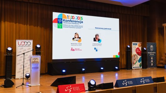 Zdjęcie na widok sali, na telebimie wyświetlani są patroni honorowi wydarzenia. Rzeczniczka Praw Dziecka i Małopolska Kurator Oświaty. 